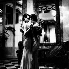 two women in evening gowns embracing each other on a checkered floor with an ornate mirror behind them