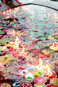 many candles are floating in the water with flowers on it and people standing around them