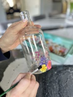 a person holding a glass bottle with flowers on it and a paintbrush in the other hand