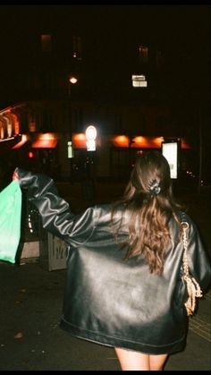 a woman in a black leather jacket is walking down the street with a green bag