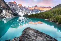 a lake surrounded by mountains and trees with blue water in the foreground at sunset