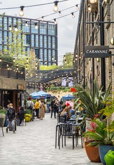 people are walking through an outdoor market with lights strung above them and plants in pots on the sidewalk