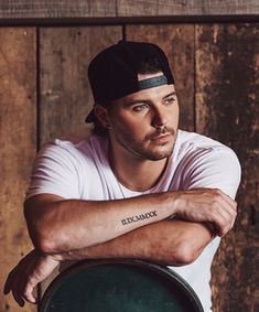 a man leaning on a barrel with his arms crossed, wearing a white shirt and black hat