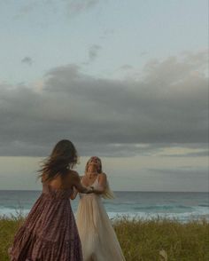 two women in long dresses are walking by the beach and one is holding her hair back