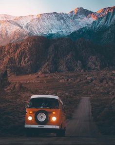 an orange and white van driving down a dirt road in front of snow capped mountains