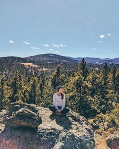 a woman sitting on top of a large rock in the middle of a pine forest