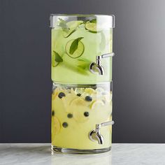 two glass containers filled with lemonade and cucumber slices on top of a marble counter