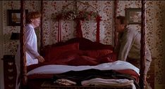 two people standing in front of a bed with red bedspread and floral wallpaper