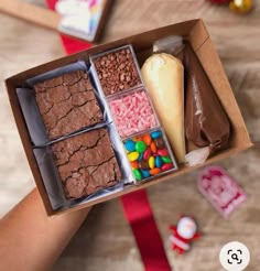 a person holding a box filled with different types of candy and brownies on top of a wooden table