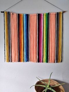 a wall hanging made with multicolored yarn next to a potted plant on a table