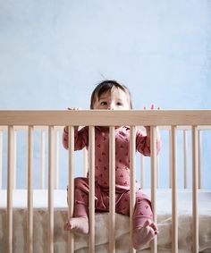 a baby sitting in a crib looking at the camera with her hands on the rails