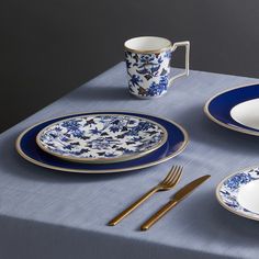 a blue and white place setting with silverware on a gray table cloth, next to a coffee cup