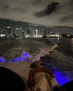 a woman laying down on the back of a boat in the water with city lights behind her