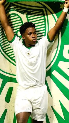 a man in white shirt and shorts standing next to a green and white wall holding up a tennis racquet