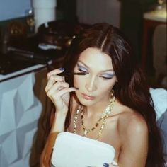 a woman with long dark hair is holding a cell phone and looking at her eyeliner