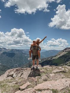 two people standing on top of a mountain with their arms in the air and one person holding an object