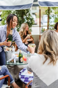 two women sitting at an outdoor table drinking wine and talking to each other while another woman holds a bottle in her hand