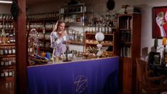 a woman standing behind a counter in a room filled with bottles and other items on shelves