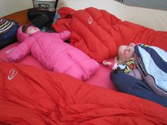 two small children sleeping on top of red sheets in a bed with pillows and blankets