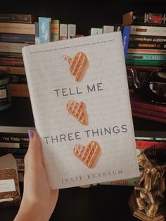 the book tell me three things is being held up in front of bookshelves