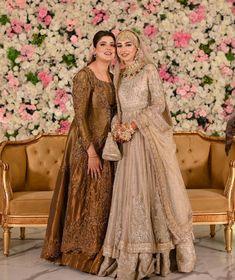 two women standing next to each other in front of a flower covered wall with flowers