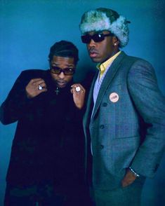 two men standing next to each other in front of a blue wall wearing sunglasses and hats