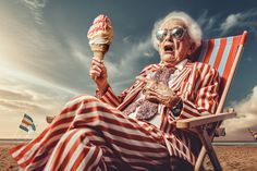 an old woman sitting in a chair on the beach eating an ice cream sundae