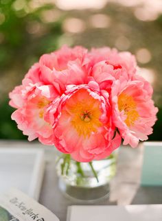 pink flowers are in a glass vase on a table next to an open book and magazine