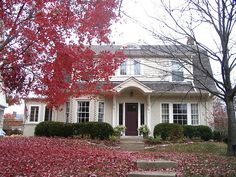 a white house with red leaves on the ground and trees in front of it,