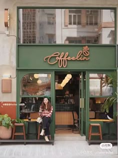 a woman sitting on a bench in front of a coffee shop with green doors and windows