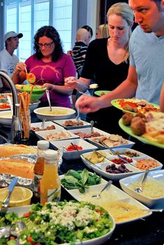 several people standing around a buffet line with food on plates and drinks in front of them