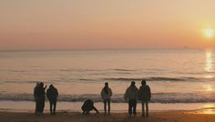 a group of people standing on top of a beach next to the ocean at sunset