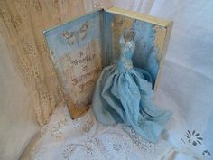 an open book with a blue dress on it next to a white lace tablecloth