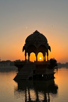 a gazebo sitting on top of a body of water at sunset with the sun in the distance