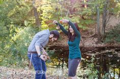 a man and woman standing next to each other in front of a pond with leaves on the ground