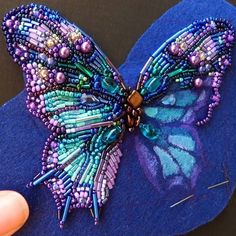 a hand holding a purple butterfly on top of a blue piece of felt with beads