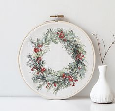 a cross stitch wreath next to a white vase with red berries and greenery on it