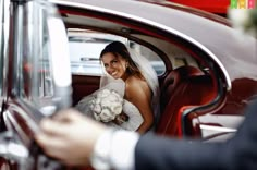 a bride sitting in the back seat of a red car with her bouquet in hand