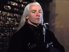 a man with long white hair wearing a black outfit and holding a microphone in his hand