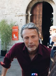 a man in a red shirt is surrounded by microphones and people outside an old building