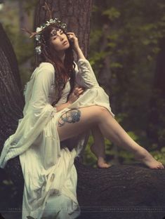 a woman sitting on top of a tree trunk wearing a white dress and flower crown