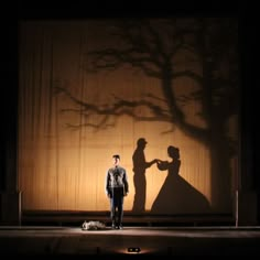 a man and woman standing on stage in front of a tree with their shadow cast