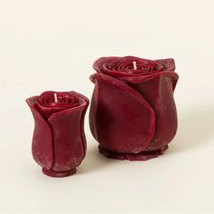 two red candles sitting next to each other on a white surface with one candle in the shape of a rose