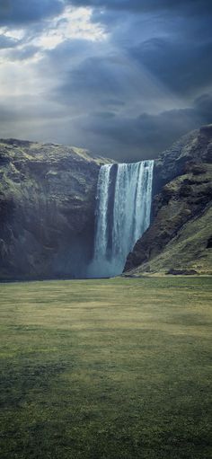 a large waterfall in the middle of a lush green field with dark clouds above it