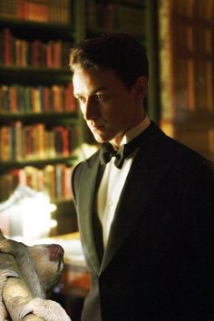 a man in a tuxedo looking down at his watch while standing next to a bookshelf