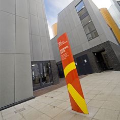 a tall orange and yellow sign sitting in front of a building next to another large gray building