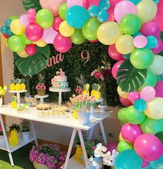 a table topped with lots of balloons and desserts next to a wall covered in greenery