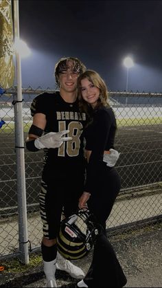 two people standing next to each other in front of a fence at night with lights on