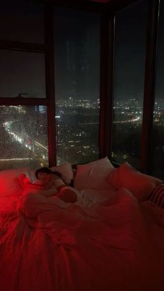 a woman is laying on her bed in front of the window at night with city lights