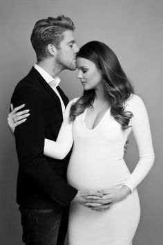 a pregnant couple kissing each other while posing for a black and white photo in front of a gray background